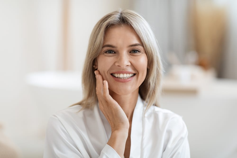 Smiling middle-aged woman touching her bright, shining skin after getting treatments to improve dullness.