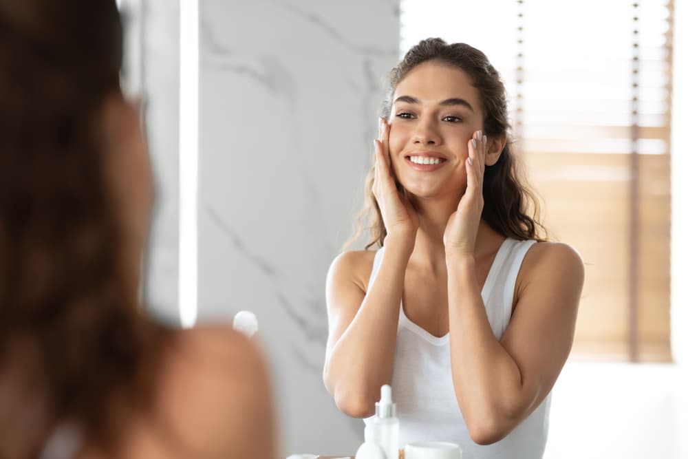 Young woman smiling and looking at her smooth skin in the mirror after getting Ultherapy skin tightening treatment.